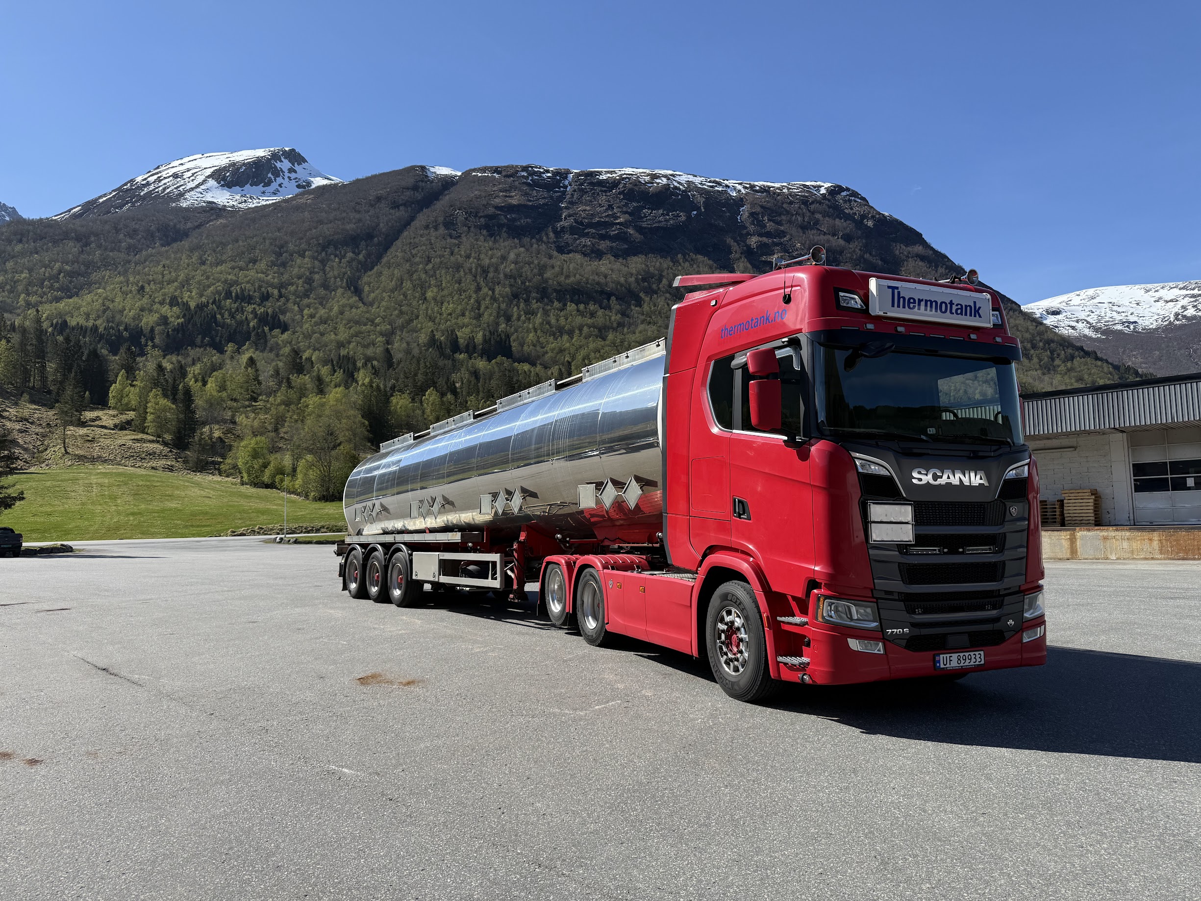 Rød tankbil klar for transportering, med nydelig natur og snøkledde fjelltopper i bakgrunnen.