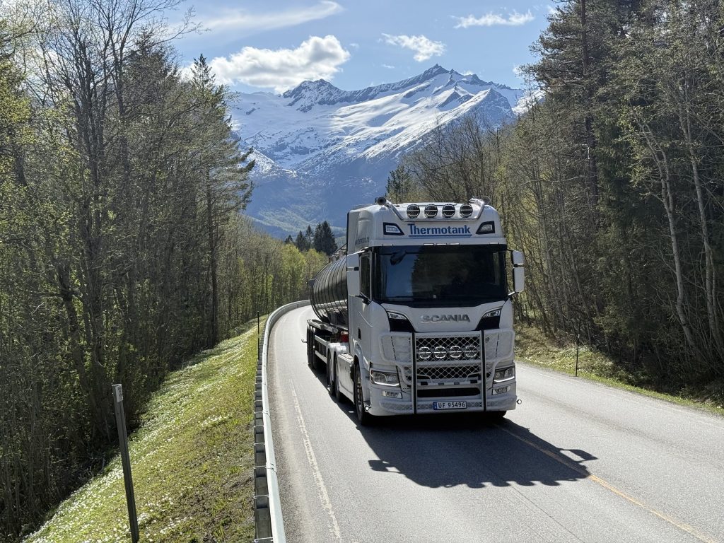 Thermotank tankbil kjører på veien med transport, med nydelig grønn natur rundt og snøkledde fjelltopper i bakgrunnen.